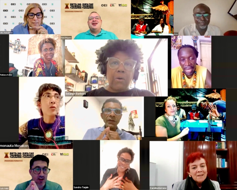 Captura de pantalla de una reunión virtual con doce participantes, cada uno de los cuales aparece en su propia ventana de vídeo. Los participantes son de diferentes edades, géneros y procedencias, y la mayoría sonríen o participan en la conversación. La pantalla incluye texto y logotipos de OEI, Trivium y «Rota dos Tambores do Atlântico» en el fondo de algunos participantes.